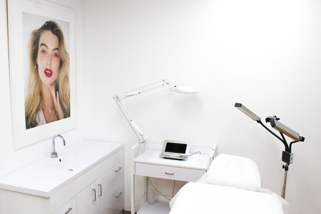Cosmetic Tattoo treatment area showing a clinic room with a framed portrait of a woman on the wall, and a trolley with a Nouveau Contour machine, with a bed and Glamcor Lights